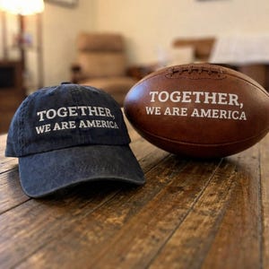 May include: A navy blue baseball cap and a brown leather football, both with the text "TOGETHER, WE ARE AMERICA" in white. The cap is slightly faded, and the football has white stitching.