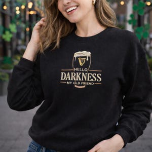 May include: Black sweatshirt with the text "HELLO DARKNESS MY OLD FRIEND" in gold and white. The design includes a beer glass graphic. The sweatshirt is a casual garment.
