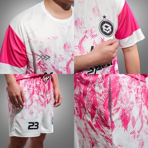 Pu&ograve; includere: Completo maglia e pantaloncini sportivi bianchi e rosa. La maglia ha maniche e dettagli rosa, con un logo circolare e la scritta "Sport" visibile. I pantaloncini hanno il numero 23. Il completo presenta un design a spruzzo.