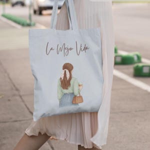 Peut inclure: Sac fourre-tout bleu clair avec le texte "La Mejor Vida" en écriture marron. Le sac présente une illustration d'une personne aux cheveux bruns, une chemise verte et une jupe bleue, tenant une tasse de café et un sac à main marron.