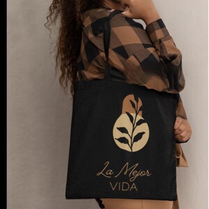 May include: Black tote bag featuring a gold-colored pear-shaped design with leaves and the text "La Mejor VIDA" in gold script. The bag is held by a person wearing a brown and black plaid shirt.