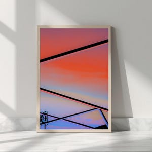 Puede incluir: Impresión artística enmarcada con un diseño abstracto con degradados de naranja, rosa y azul. Líneas negras se cruzan sobre el fondo colorido, creando formas geométricas. La obra de arte se presenta en un marco de madera clara.