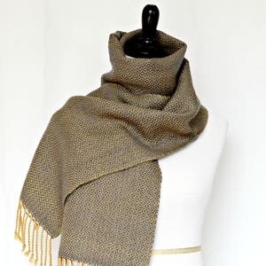 May include: A woven scarf with a geometric pattern in shades of gray and gold. The scarf has a fringe on both ends.