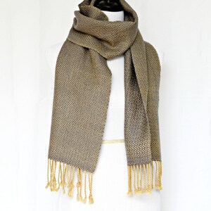 May include: A gray and gold woven scarf with a geometric pattern and fringe. The scarf is displayed on a white mannequin.