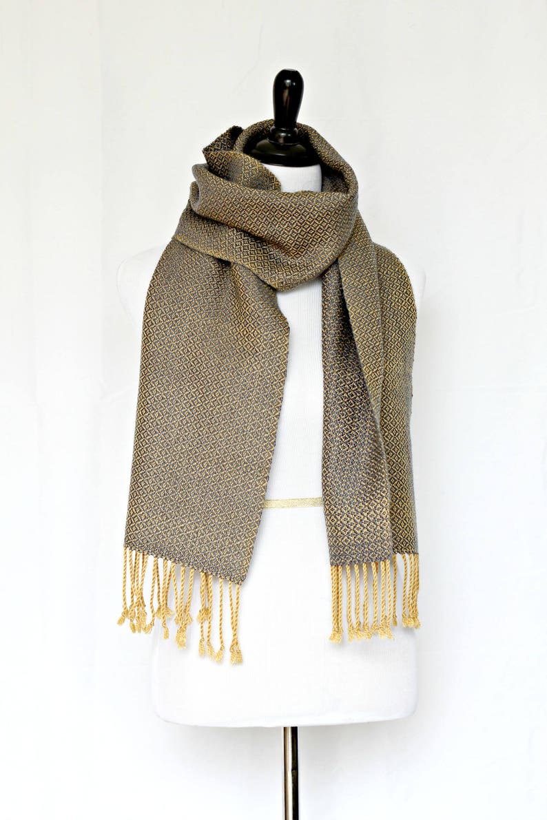 May include: A gray and gold woven scarf with a geometric pattern and fringe. The scarf is displayed on a white mannequin.