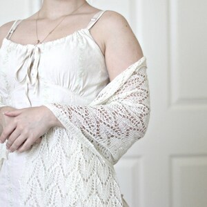 May include: A white crocheted shawl with a delicate lace pattern. The shawl is draped over a person's shoulders and arms, and the person is wearing a white sleeveless dress.