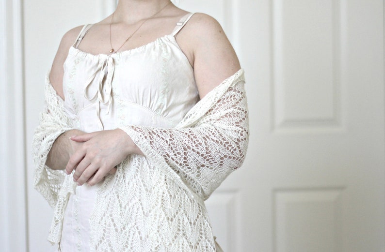 May include: A white crocheted shawl with a delicate lace pattern. The shawl is draped over a person's shoulders and arms, and the person is wearing a white sleeveless dress.