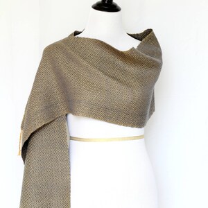 May include: A gray and gold woven scarf with a geometric pattern and fringe. The scarf is draped over a white mannequin.
