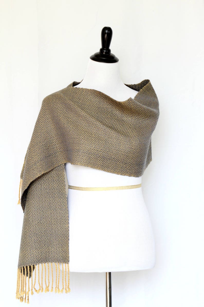 May include: A gray and gold woven scarf with a geometric pattern and fringe. The scarf is draped over a white mannequin.