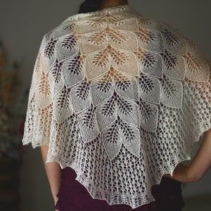 May include: A white crocheted shawl with a delicate leaf pattern. The shawl is draped over a person's shoulders and is made of a fine, lightweight yarn.
