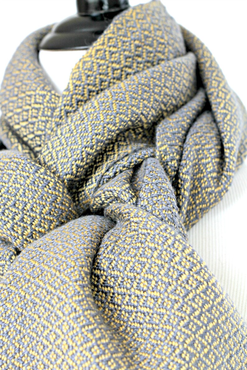 May include: A gray and yellow woven scarf with a geometric pattern. The scarf is folded and draped over a mannequin.