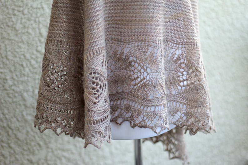 May include: A delicate, beige knitted shawl with a lacy, openwork design. The shawl is draped over a mannequin and has a scalloped edge.