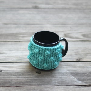 May include: A black mug with a teal knitted cosy. The cosy has a textured pattern of small bumps.