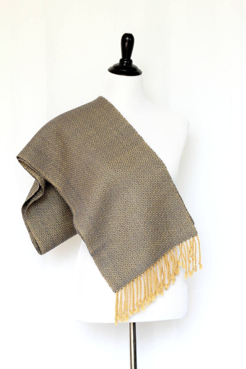 May include: A gray and gold woven scarf with a geometric pattern and gold fringe.