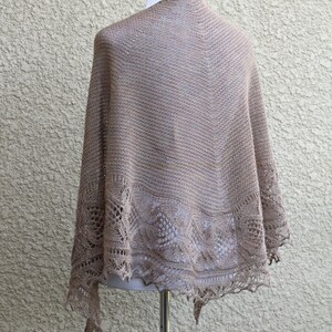 May include: A beige knitted shawl with a delicate lace pattern around the edges. The shawl is draped over a mannequin.