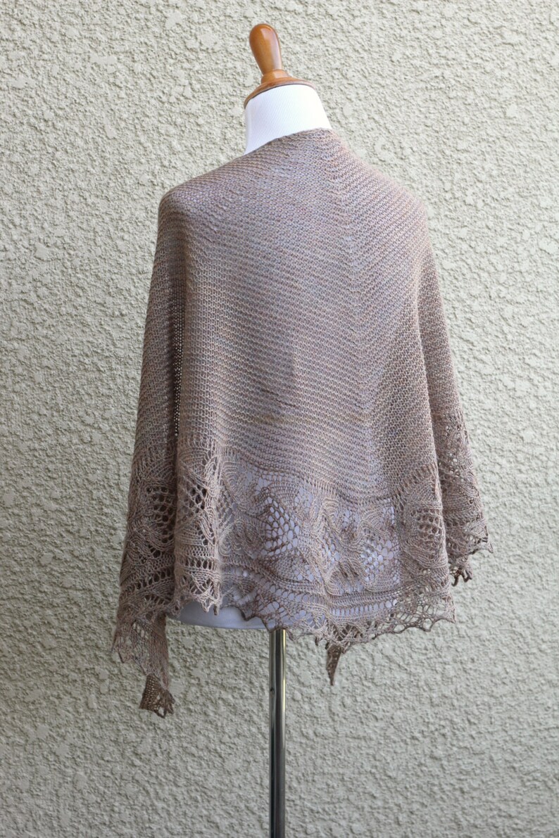May include: A beige knitted shawl with a delicate lace pattern around the edges. The shawl is draped over a mannequin.