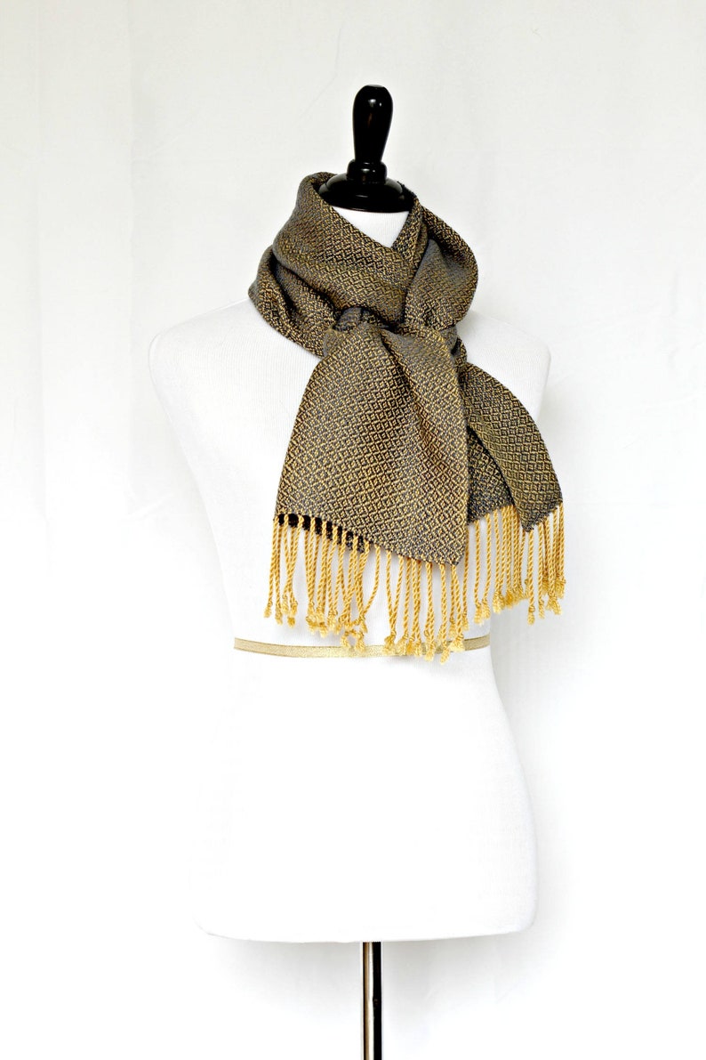 May include: A gray and gold patterned scarf with gold fringe. The scarf is tied around a white mannequin.