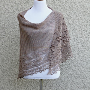May include: A brown knitted shawl with a delicate lace pattern, displayed on a white mannequin against a textured beige wall.