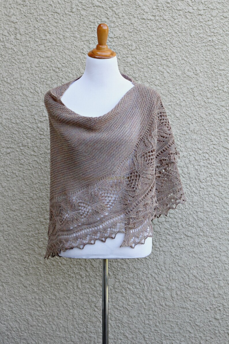 May include: A brown knitted shawl with a delicate lace pattern, displayed on a white mannequin against a textured beige wall.