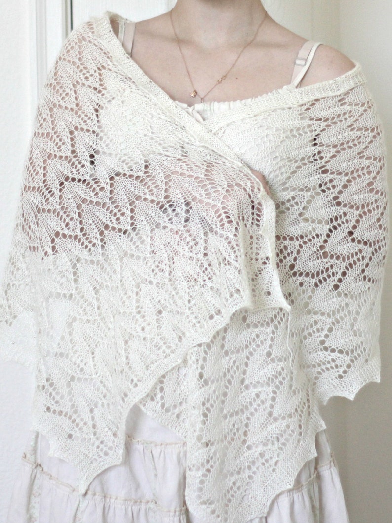 May include: A white crocheted shawl with a delicate lace pattern. The shawl is draped over a person's shoulders and is worn with a white dress.