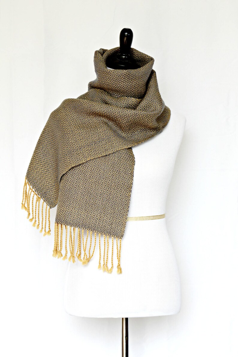 May include: A woven scarf with a geometric pattern in shades of gray and gold. The scarf has a fringe on both ends.