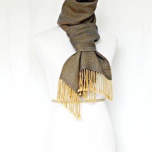 May include: A gray and gold woven scarf with fringe. The scarf is displayed on a white mannequin.