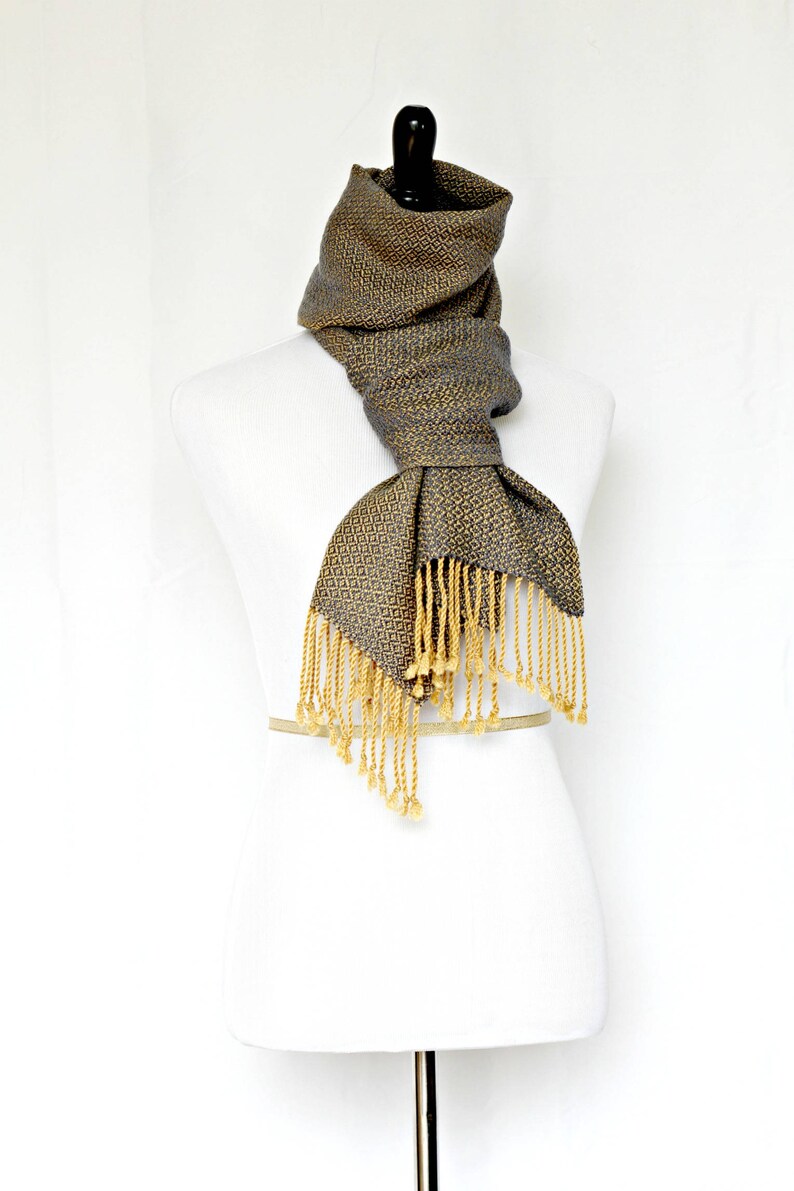 May include: A gray and gold woven scarf with fringe. The scarf is displayed on a white mannequin.