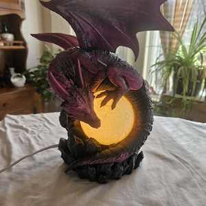 May include: A purple dragon lamp with a glowing orb. The dragon is perched atop a rocky base, with its wings spread. The lamp is plugged in with a gray cord. The dragon's wings and body have a textured appearance.