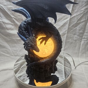 May include: A detailed dragon figurine in shades of blue and black, coiled around a glowing, yellow moon. The dragon's wings are spread, and the sculpture sits on a mirrored base. The figurine is lit from within, creating a fantasy-themed decorative piece.