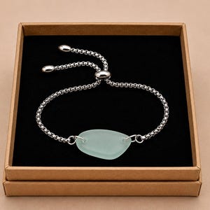 May include: A silver-toned adjustable bracelet with a light blue, oval-shaped sea glass centerpiece. The bracelet is presented in a brown cardboard box with a black interior, set against a neutral background.