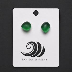 Puede incluir: Un par de pendientes de botón de cristal de mar verde esmeralda, pequeños, lisos y redondeados, sujetos a una tarjeta de presentación rectangular blanca. La tarjeta presenta un gráfico negro de una ola estilizada y el texto FAVIERI JEWELRY.