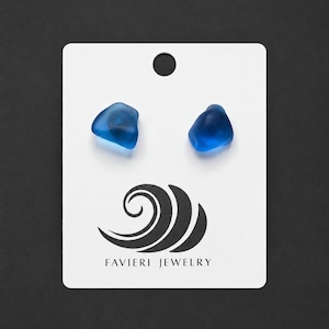 Peut inclure: Une paire de boucles d'oreilles à clous en verre de mer bleu cobalt fixées sur une carte de présentation rectangulaire blanche. La carte présente un motif noir de vague stylisée et le texte FAVIERI JEWELRY imprimé en noir en bas.