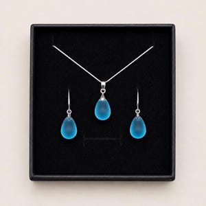 Puede incluir: Un conjunto de joyas que presenta un collar de plata y pendientes a juego, presentado en una caja negra. El conjunto incluye un colgante y dos pendientes en forma de lágrima, todos en un color azul claro translúcido.