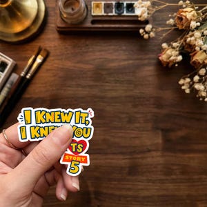 Puede incluir: Una pegatina de vinilo troquelada con el texto amarillo I KNEW IT, I KNEW YOU, un coraz&oacute;n rojo y el texto azul y naranja TOY STORY 5. La pegatina tiene un borde blanco y se sostiene sobre un fondo de madera oscura con material de arte y flores secas.