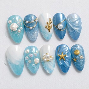 May include: A set of ten artificial nails with a mermaid theme. The nails feature various designs, including glitter, seashells, pearls, and gold accents. The color palette consists of shades of blue and white, creating a coastal aesthetic.
