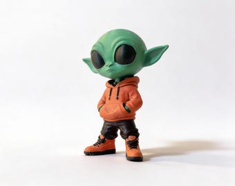 Puede incluir: Una figura de alienígena verde con grandes ojos negros, que lleva una sudadera con capucha naranja, pantalones negros y botas naranjas. La figura tiene las manos en los bolsillos y está de pie sobre una superficie blanca. La figura es una representación estilizada de un alienígena.
