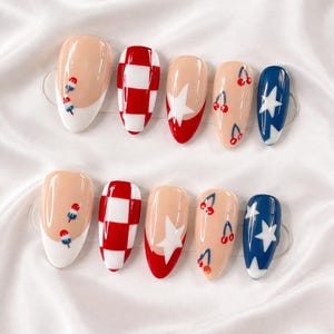 May include: A set of ten artificial nails with various designs. The designs include red and white checkerboard, white stars on a blue background, cherries, and popsicle-like shapes. The nails have a glossy finish and are arranged on a white surface.