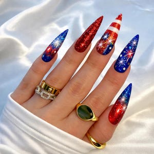 May include: A hand with long, pointed fingernails decorated with a patriotic theme. The nails feature designs of fireworks, glitter, and stripes in red, white, and blue. Gold rings adorn the fingers.