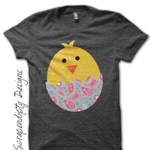 May include: A dark gray t-shirt with a yellow chick peeking out of a blue, pink, and green patterned egg.
