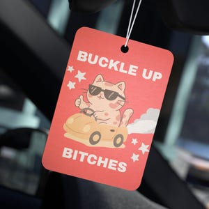 May include: A red rectangular car air freshener with the text "BUCKLE UP BITCHES". The air freshener features a cartoon cat wearing sunglasses and driving a yellow car, with white stars around the image.