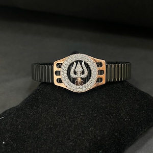 Puede incluir: Una pulsera negra y dorada con una pieza central circular. La pieza central presenta un diseño de tridente rodeado de pequeñas piedras brillantes. La pulsera tiene una textura acanalada.