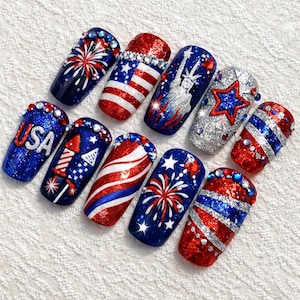 May include: A set of ten artificial nails with patriotic designs. The nails feature red, white, and blue glitter, stripes, stars, fireworks, and the Statue of Liberty. Some nails have the word "USA" in red.