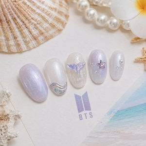 May include: A set of five press-on nails featuring a pearlescent white and lavender shimmer finish. The designs include a silver wave accent, a three-dimensional purple mermaid tail, a small purple star charm, and tiny white pearls.
