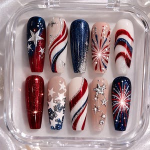 May include: A set of ten artificial nails with patriotic designs. The nails feature red, white, and blue colors with glitter, stars, and firework patterns. The nails are displayed in a clear plastic case.