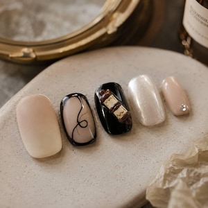 May include: A set of five press-on nails featuring a variety of designs: one solid nude, one nude with a black line accent, one black with a 3D scroll charm, one pearlescent white, and one nude with a single small clear rhinestone accent.