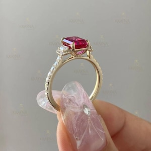 Könnte beinhalten: Ein goldener Ring mit einem großen, rechteckigen, roten Edelstein. Der Ring hat einen Halo aus kleineren, klaren Edelsteinen um den Hauptstein und entlang des Bandes. Der Ring wird hochgehalten, um sein Design und seinen Glanz zu zeigen.