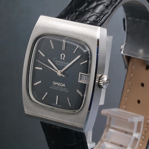 Könnte beinhalten: Eine Vintage Omega Constellation Armbanduhr mit einem dunkelgrauen quadratischen Zifferblatt und silbernen Zeigern und Markierungen. Die Uhr hat ein Edelstahlgehäuse und ein schwarzes Lederarmband. Das Datum wird bei 3 Uhr angezeigt.