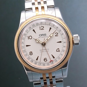 Może przedstawiać: Srebrno-złoty zegarek na rękę z białą tarczą. Zegarek posiada markę "ORIS" oraz napisy "25 JEWELS" i "AUTOMATIC SWISS MADE". Zegarek ma wyświetlacz daty i srebrno-złotą bransoletę.