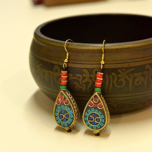 Può includere: Un paio di orecchini pendenti color oro con un ciondolo a forma di goccia. Il ciondolo presenta un design a mosaico in blu, rosso e oro, con un accento di perline rosse e verdi. Gli orecchini sono esposti vicino a una ciotola decorativa.