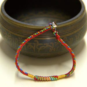May include: A vibrant braided bracelet featuring red, yellow, blue, and green threads. The bracelet has a small silver-coloured bell at the top and gold-coloured accents. The bracelet is likely handmade and is set against a blurred background.
